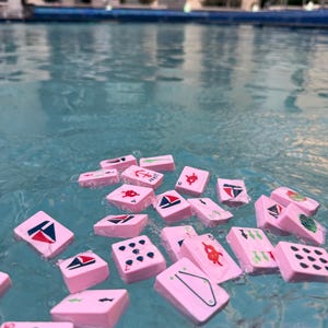 May include: Pink Mahjong tiles floating in a pool. The tiles feature various red and white nautical-themed designs, including sailboats, anchors, and geometric shapes. The water is a clear blue, reflecting the surrounding buildings.