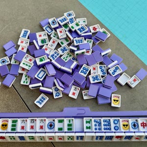 May include: A Mahjong set with purple tiles and a matching rack. The tiles are white with various symbols and characters, including Jokers with sunglasses. The set is arranged on a gray surface with a turquoise background.