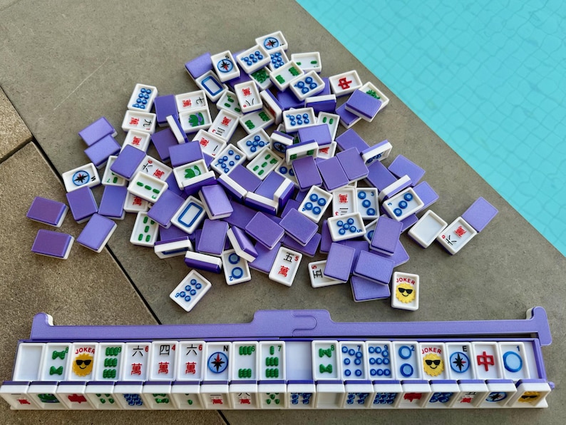 May include: A Mahjong set with purple tiles and a matching rack. The tiles are white with various symbols and characters, including Jokers with sunglasses. The set is arranged on a gray surface with a turquoise background.