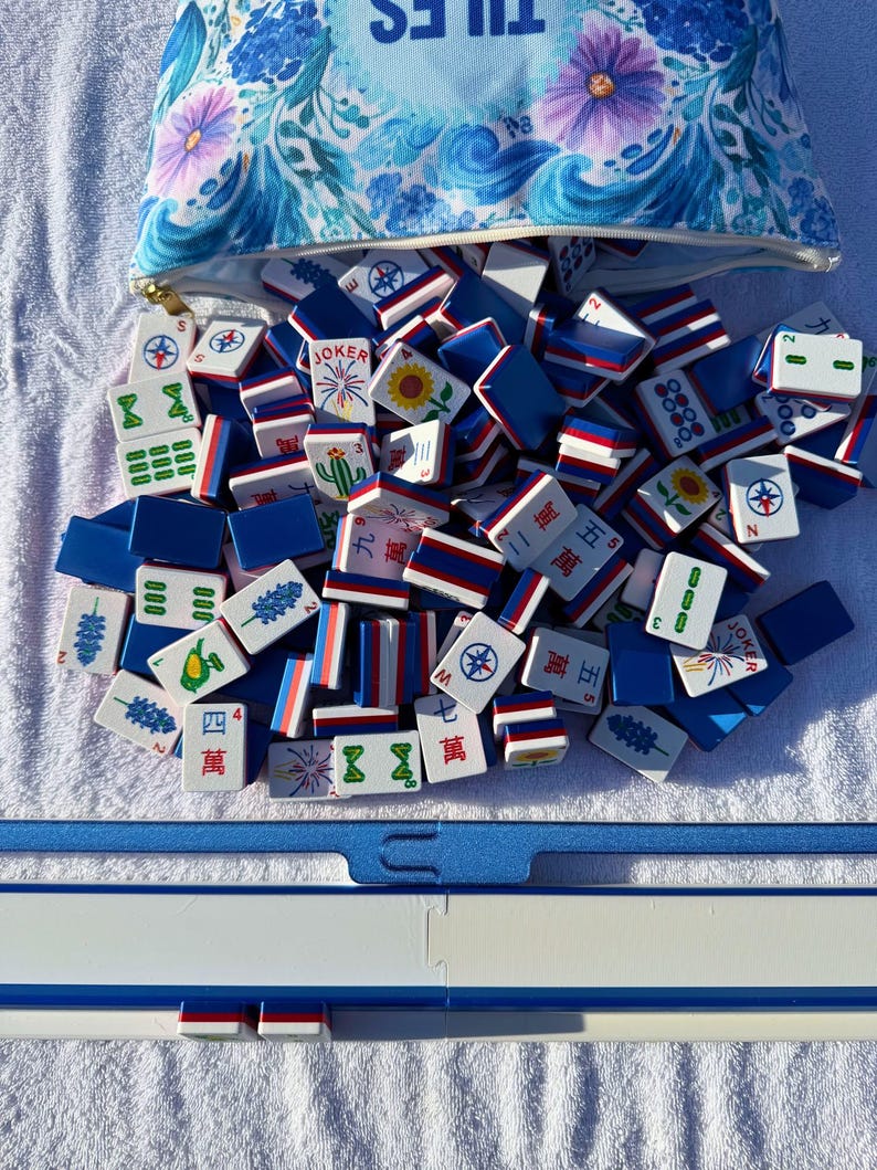 May include: A collection of Mahjong tiles spilling from a blue floral patterned bag. The tiles are white, blue, and red, with various symbols and Chinese characters. A blue Mahjong table is visible at the bottom.