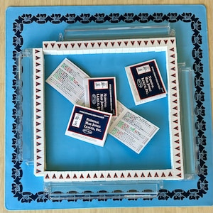 Könnte beinhalten: Ein komplettes Mahjongg-Set mit weißen Spielsteinen, roten und schwarzen Markierungen und transparenten Kunststoffgestellen, angeordnet auf einer blauen Matte mit floralem Rand. Mehrere Karten mit dem Text "NATIONAL MAH JONGG LEAGUE, INC." liegen herum.
