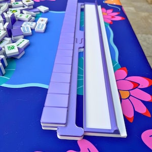 May include: A purple Mahjong tile rack and tiles are displayed on a blue surface with a floral design. The tiles are white with purple markings. The rack is made of purple plastic.