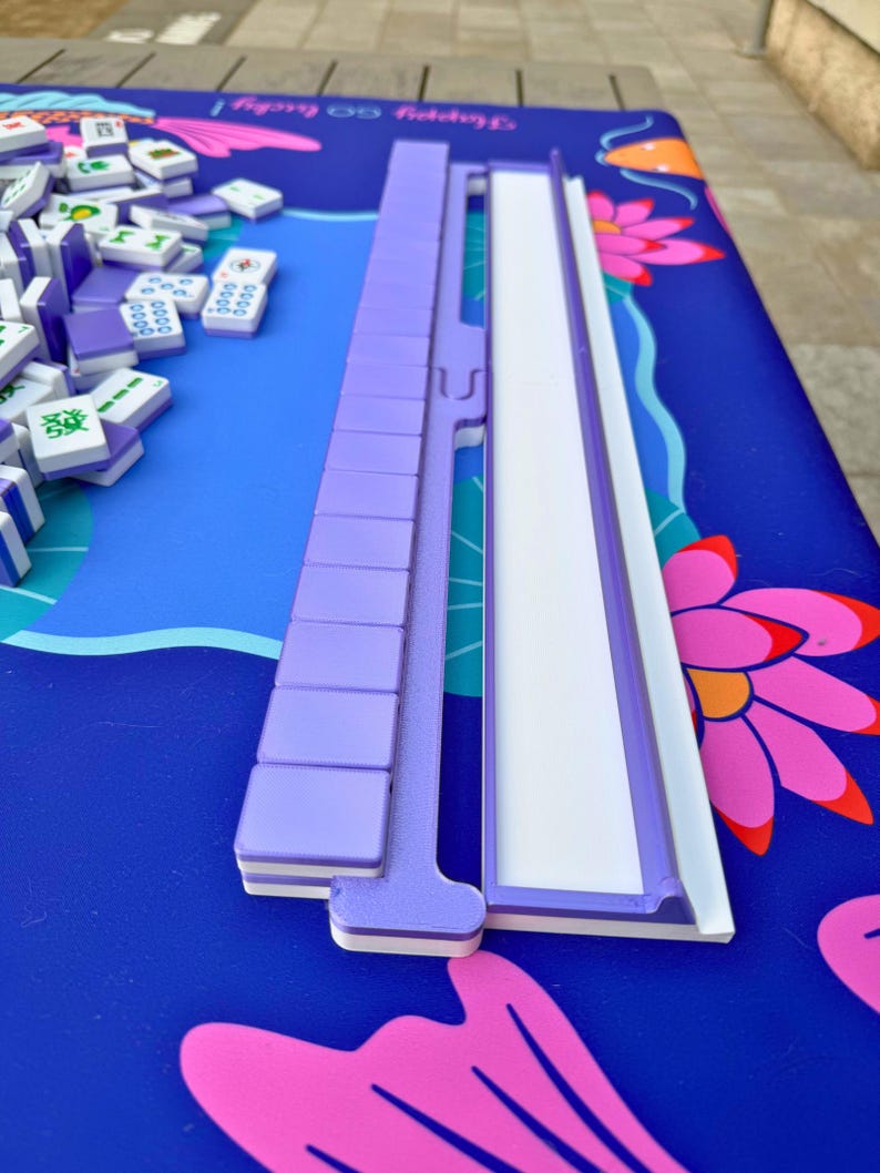 May include: A purple Mahjong tile rack and tiles sit on a blue surface with a floral design. The tiles are white with purple markings. The rack is made of purple plastic and has a white inner track.
