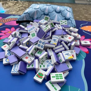May include: A pile of Mahjong tiles with purple backs and white faces, scattered on a blue surface with a floral pattern. The tiles display various symbols and numbers, including the word "Joker" and Chinese characters.