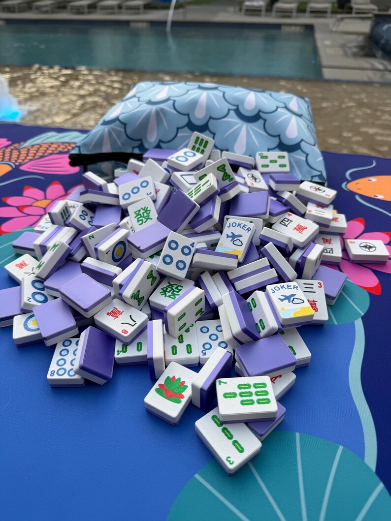 May include: A pile of Mahjong tiles with purple backs and white faces, scattered on a blue surface with a floral pattern. The tiles display various symbols and numbers, including the word "Joker" and Chinese characters.
