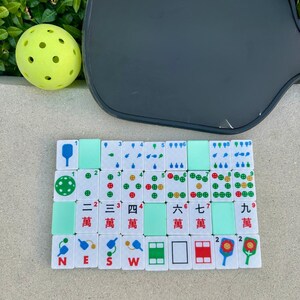 May include: A collection of white Mahjong tiles with green and red markings, arranged on a surface. A yellow pickleball and a dark gray pickleball paddle are also in the image. The tiles display Chinese characters and symbols.