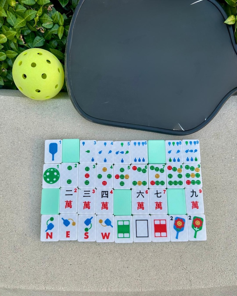 May include: A collection of white Mahjong tiles with green and red markings, arranged on a surface. A yellow pickleball and a dark gray pickleball paddle are also in the image. The tiles display Chinese characters and symbols.