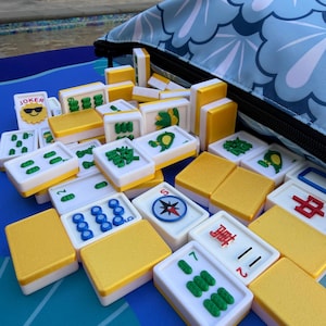 May include: A collection of Mahjong tiles with white faces, green and blue symbols, and yellow backs. The tiles are scattered on a blue surface next to a blue floral patterned bag. One tile reads "JOKER".