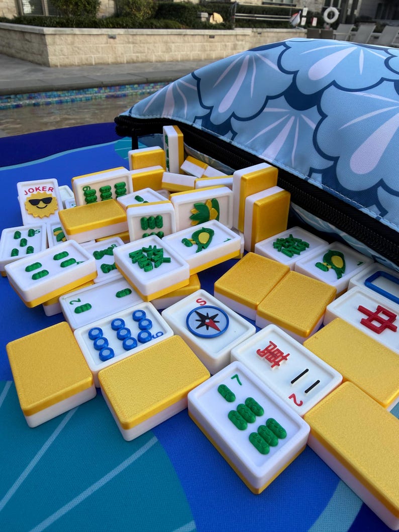 May include: A collection of Mahjong tiles with white faces, green and blue symbols, and yellow backs. The tiles are scattered on a blue surface next to a blue floral patterned bag. One tile reads "JOKER".
