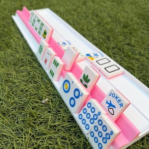 May include: A white and pink Mahjong set with tiles displaying blue and green symbols and numbers. The tiles are arranged in a pink channel on a white tray. Some tiles have the word "Joker" printed on them.