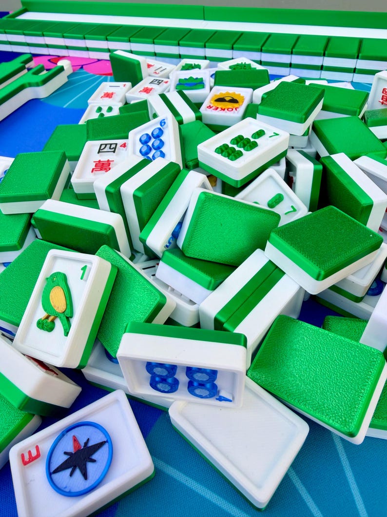 May include: A collection of green and white Mahjong tiles, some with symbols and numbers, others with green textured surfaces. The tiles are scattered on a blue surface, with some arranged in rows in the background. The game pieces are a mix of shapes and designs.
