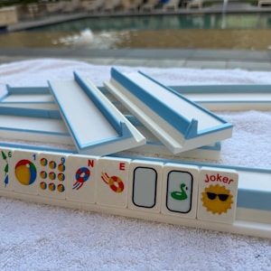 May include: A Mahjong set with white tiles featuring colorful symbols and characters, including a sun with sunglasses and the word "Joker." The set includes blue and white racks.