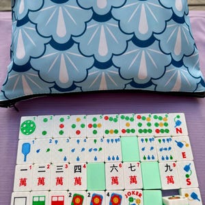 May include: A Mahjong set with white tiles featuring Chinese characters and colorful symbols. The set includes green tiles and a light blue carrying case with a dark blue and white fan-like pattern. The tiles are arranged on a purple surface.