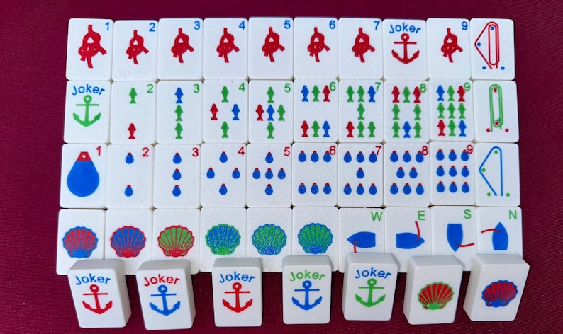May include: A complete set of Mahjong tiles, featuring white tiles with red, green, and blue symbols. The set includes numbered tiles, Jokers with anchors, and directional wind tiles. The tiles are arranged on a burgundy surface.