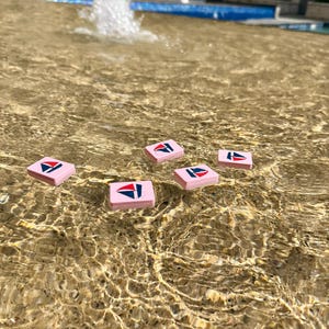May include: Six pink rectangular tiles with a sailboat logo in red, white, and blue float in a fountain. The water's surface reflects sunlight, creating ripples around the tiles. The tiles are evenly spaced in the water.