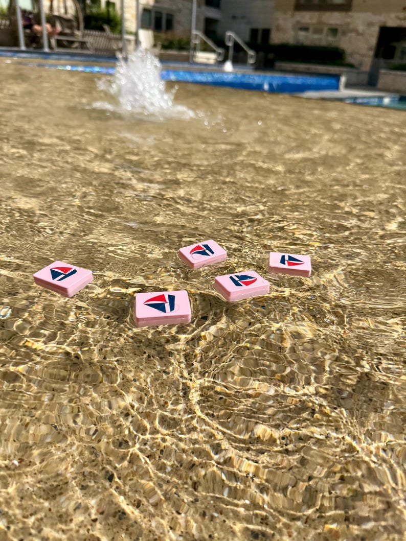 May include: Six pink rectangular tiles with a sailboat logo in red, white, and blue float in a fountain. The water's surface reflects sunlight, creating ripples around the tiles. The tiles are evenly spaced in the water.