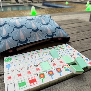 May include: A Mahjong set with white tiles featuring colorful symbols and Chinese characters. The set includes green tiles and is displayed on a wooden surface. A blue pouch with a white and blue pattern is in the background.