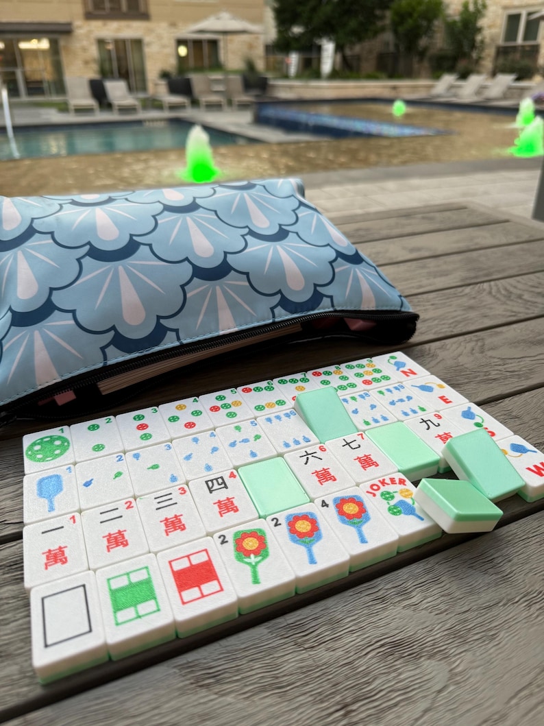May include: A Mahjong set with white tiles featuring colorful symbols and Chinese characters. The set includes green tiles and is displayed on a wooden surface. A blue pouch with a white and blue pattern is in the background.