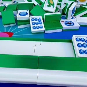 May include: A close-up shot of a Mahjong game in progress. The tiles are white with green backs and feature various symbols and numbers. Some tiles have a bird illustration. The game is set on a blue and white patterned surface.