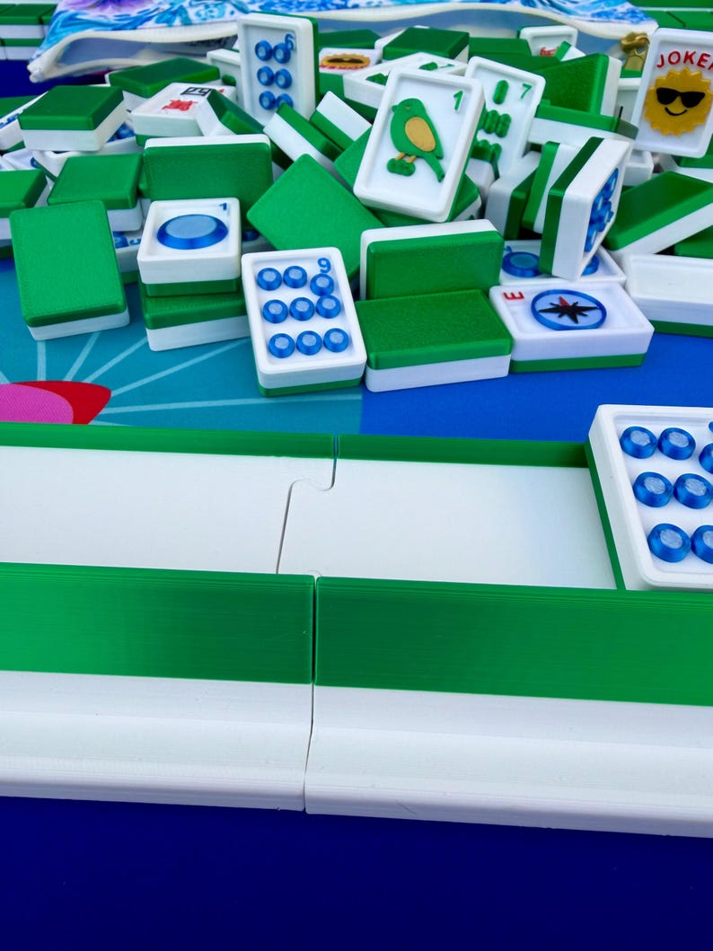 May include: A close-up shot of a Mahjong game in progress. The tiles are white with green backs and feature various symbols and numbers. Some tiles have a bird illustration. The game is set on a blue and white patterned surface.