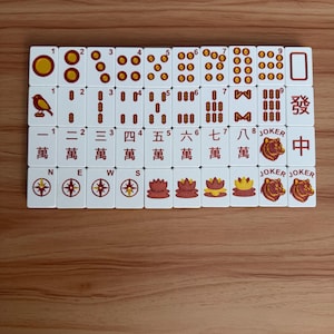 May include: A set of white Mahjong tiles with red and gold markings, arranged on a wooden surface. The tiles feature various symbols, numbers, and Chinese characters. Some tiles have Jokers and compass rose designs.