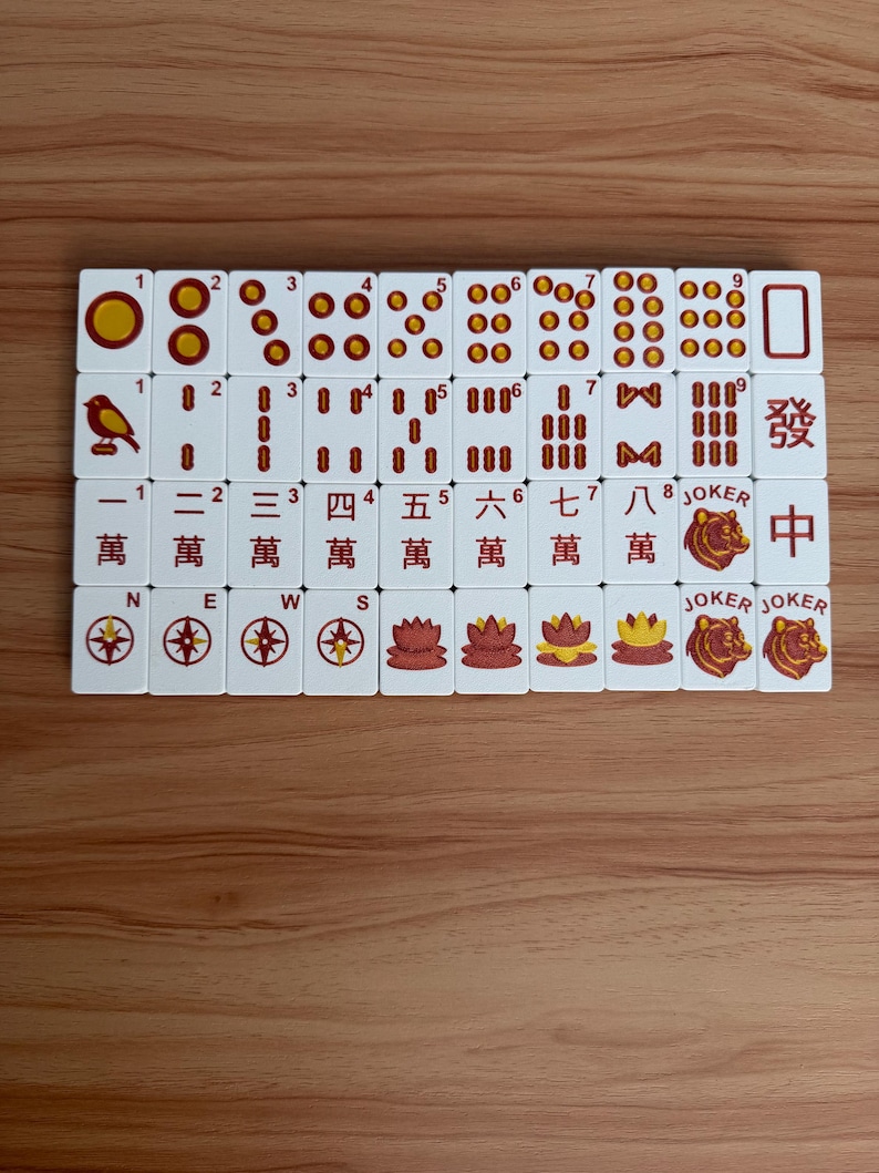May include: A set of white Mahjong tiles with red and gold markings, arranged on a wooden surface. The tiles feature various symbols, numbers, and Chinese characters. Some tiles have Jokers and compass rose designs.