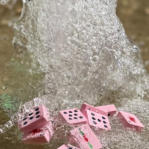 May include: Pink dominoes with black dots and red and green symbols are partially submerged in water, with a splash of water in the background. The dominoes are scattered, creating a playful and dynamic scene. The water appears clear and clean.