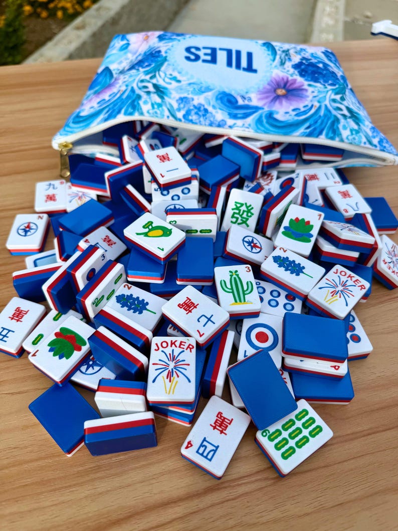 May include: A collection of Mahjong tiles in a blue and white floral patterned pouch. The tiles are white with blue and red accents, featuring various symbols and characters. The pouch has the word "TILES" printed on it.
