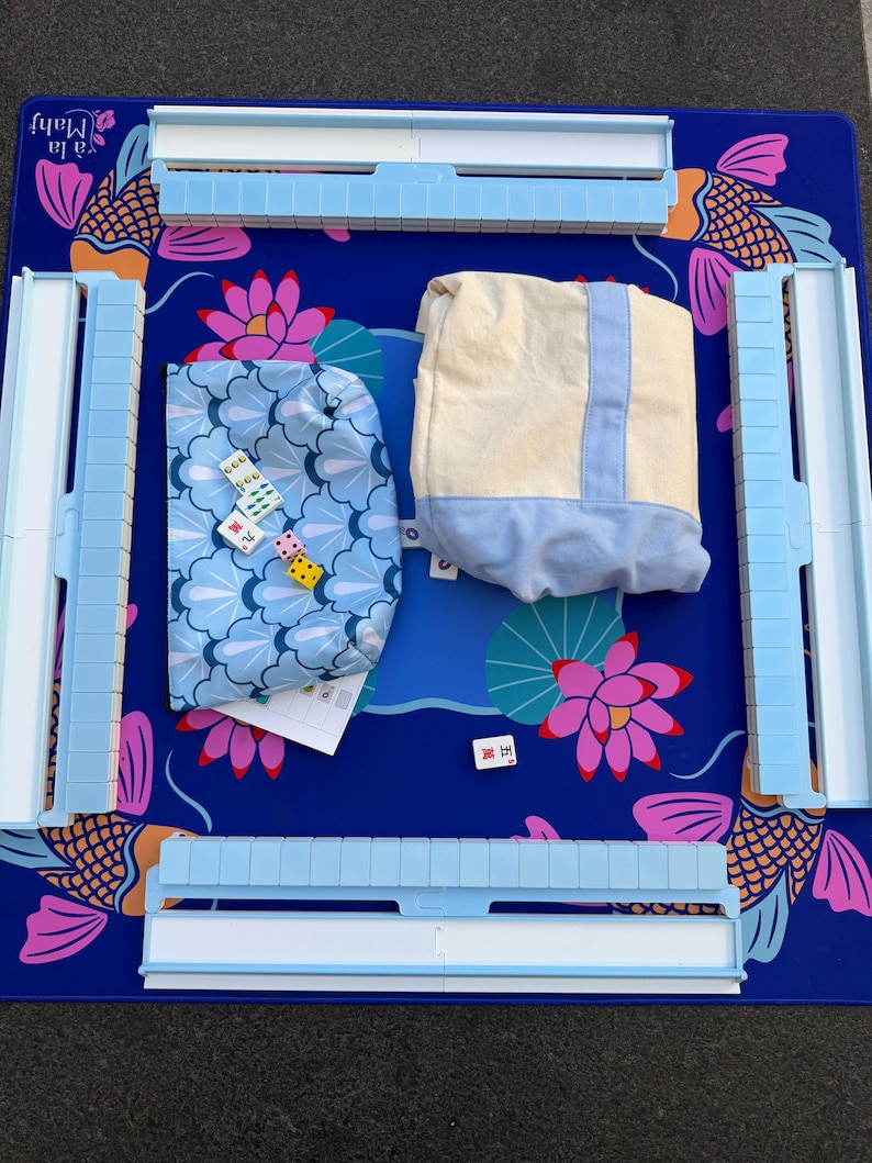 May include: A Mahjong set with a blue playing surface featuring koi fish and lotus flower designs. The set includes light blue tile racks, two fabric pouches, dice, and tiles. The game board has the text "Ya la Mahf" in the upper left corner.
