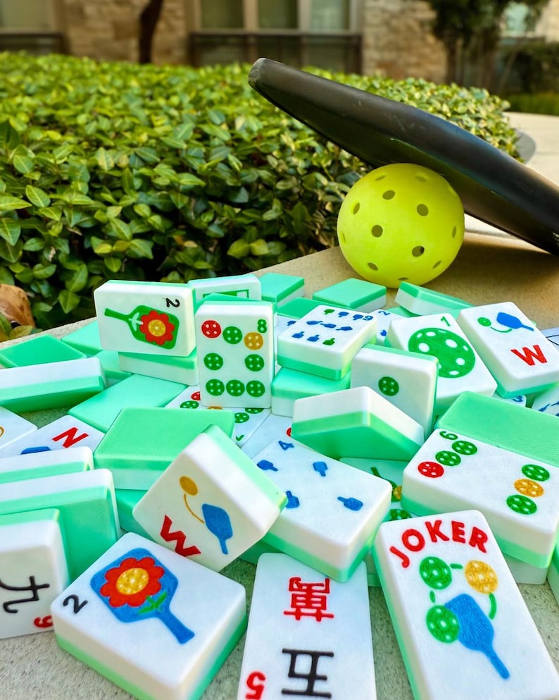 May include: A collection of white and green Mahjong tiles with various symbols and numbers. A yellow pickleball and a black paddle are in the background. The tiles are arranged on a light-colored surface.