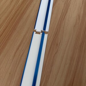 May include: Two white and blue plastic pieces, possibly for a track or guide, are shown against a wood grain background. The pieces have a white center, blue edges, and a connecting notch. The design is simple and functional.