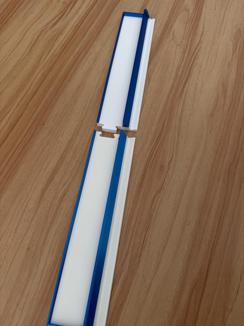 May include: Two white and blue plastic pieces, possibly for a track or guide, are shown against a wood grain background. The pieces have a white center, blue edges, and a connecting notch. The design is simple and functional.