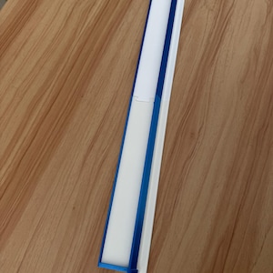 May include: A long, rectangular object with a white interior and a blue frame. The object is made of plastic and has a hinged design. The object is lying on a wooden surface.