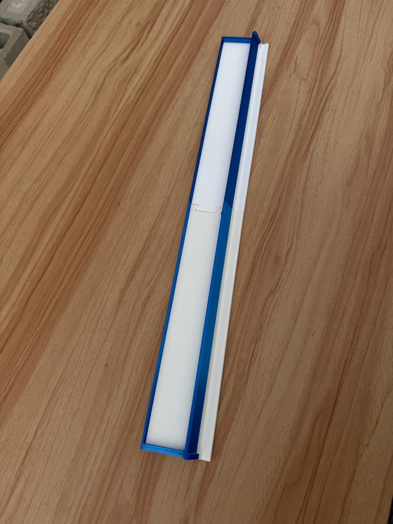 May include: A long, rectangular object with a white interior and a blue frame. The object is made of plastic and has a hinged design. The object is lying on a wooden surface.