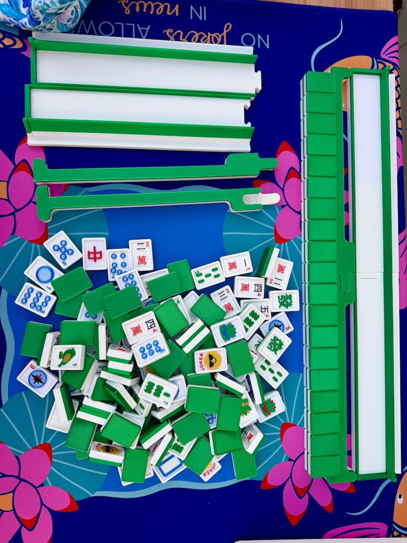 May include: A complete Mahjong set with green and white components, including tiles and racks, arranged on a blue surface with floral accents. The tiles feature various symbols and characters. The text "NO Jokers ALLOW IN news" is visible.