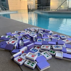 May include: A collection of purple and white Mahjong tiles scattered on a gray surface. The tiles feature various symbols and numbers in red, green, and blue. The background includes a swimming pool and tall buildings.