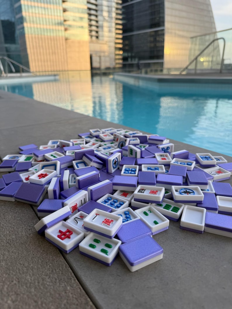May include: A collection of purple and white Mahjong tiles scattered on a gray surface. The tiles feature various symbols and numbers in red, green, and blue. The background includes a swimming pool and tall buildings.