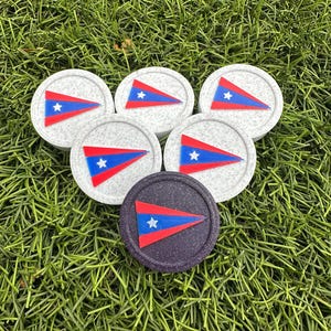 May include: A collection of round coasters with a flag design. The coasters are white and dark purple with a red, blue, and white flag emblem. The coasters are arranged on a bed of green grass.