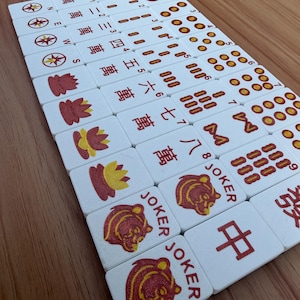 May include: A collection of white Mahjong tiles with red and yellow symbols and Chinese characters. The tiles are arranged on a wooden surface, showcasing various suits and joker tiles.