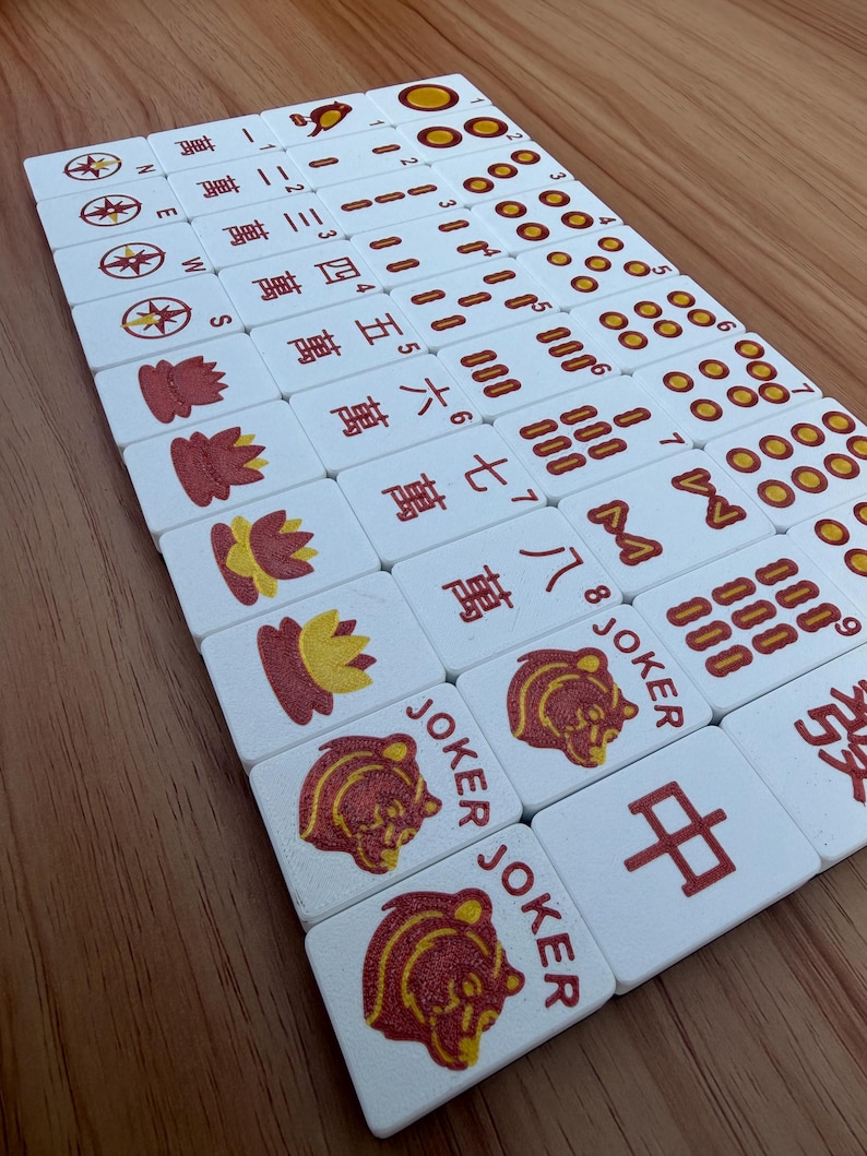 May include: A collection of white Mahjong tiles with red and yellow symbols and Chinese characters. The tiles are arranged on a wooden surface, showcasing various suits and joker tiles.