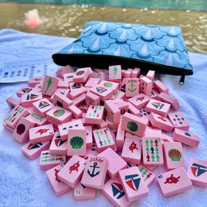 May include: A pink Mahjong set with various nautical-themed symbols, including anchors, sailboats, and shells. The set is accompanied by a blue pouch with a wave pattern. The game is set on a white towel near a pool.