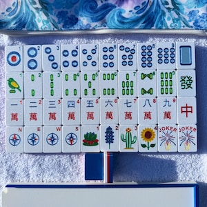 May include: A complete Mahjong set with white tiles featuring various symbols and numbers. The set includes tiles with dots, bamboo, characters, and Jokers. A blue floral patterned case and a white and blue tile rack are also visible.