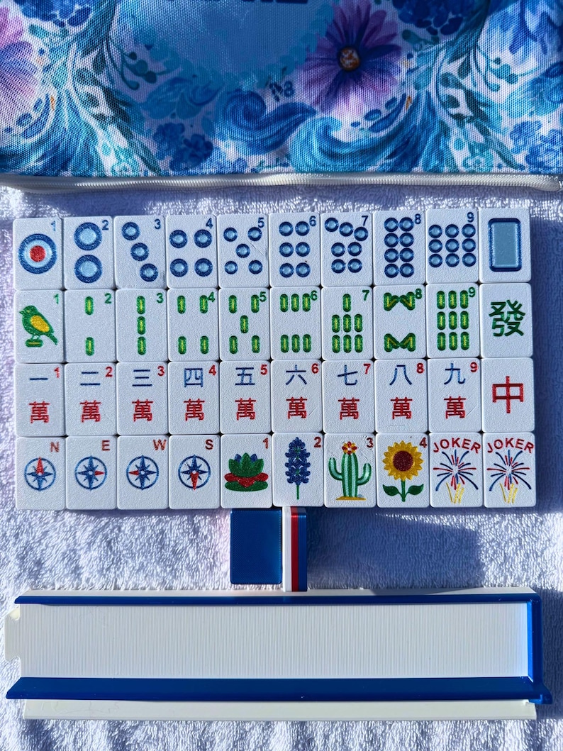 May include: A complete Mahjong set with white tiles featuring various symbols and numbers. The set includes tiles with dots, bamboo, characters, and Jokers. A blue floral patterned case and a white and blue tile rack are also visible.