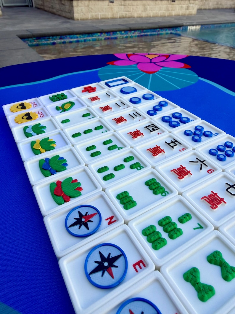 May include: A set of white Mahjong tiles with colorful symbols and characters, including compass rose designs and green, yellow, and red decorative elements. The tiles are arranged on a blue surface with a floral pattern.
