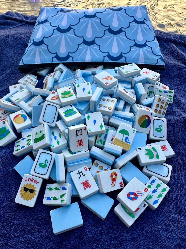 May include: A collection of Mahjong tiles with various symbols and numbers, scattered on a blue surface. A matching blue patterned bag is in the background. The tiles are white with blue accents and feature illustrations and Chinese characters.