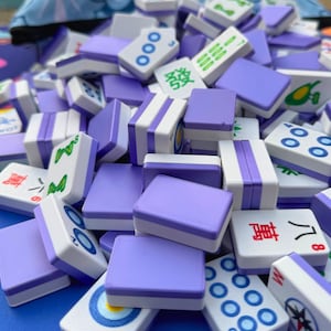 May include: A pile of Mahjong tiles in shades of purple and white. The tiles feature various symbols and characters, including circles, lines, and Chinese characters. The tiles are stacked, creating a visually appealing arrangement.