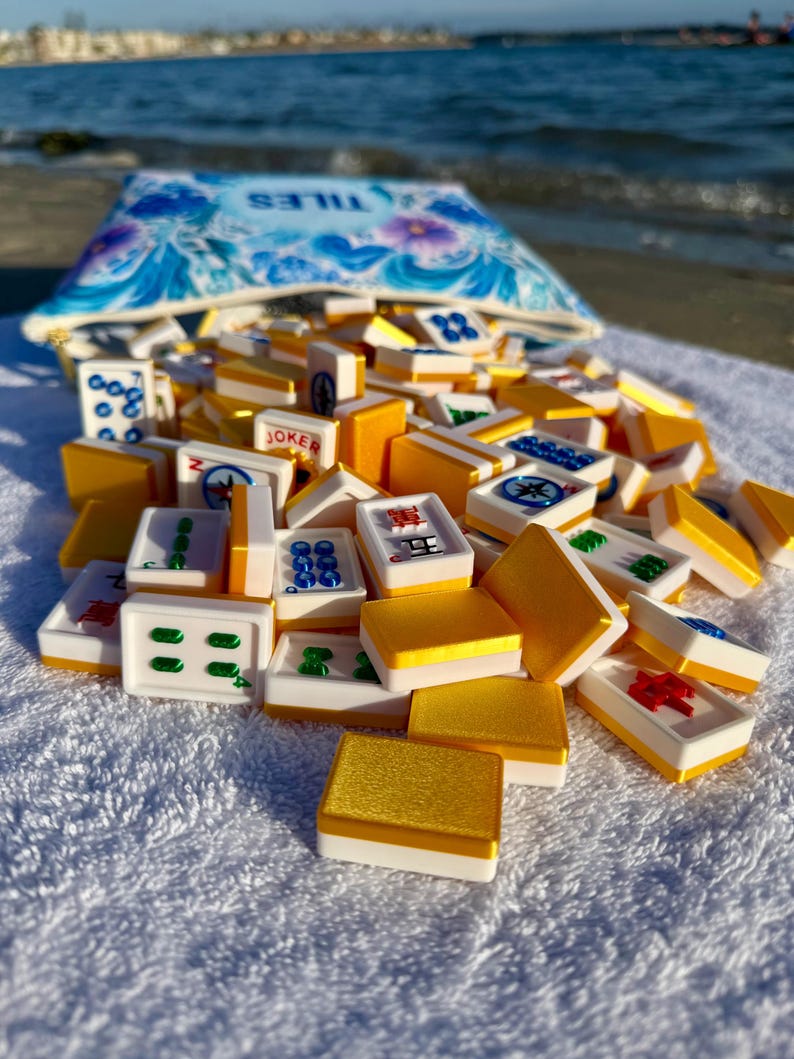 May include: A collection of Mahjong tiles spilling from a blue floral patterned pouch. The tiles are white with various symbols and numbers, and gold and yellow accents. The pouch has the word "TILES" printed on it.