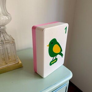 May include: A white, rectangular decorative box with a pink edge and a green bird illustration with a yellow belly. The number "1" is in the upper right corner. The box is on a light blue table.