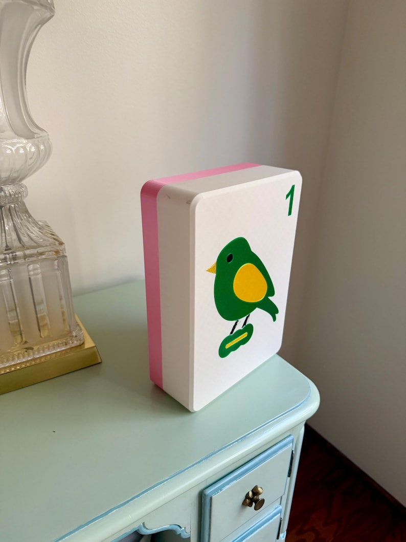 May include: A white, rectangular decorative box with a pink edge and a green bird illustration with a yellow belly. The number "1" is in the upper right corner. The box is on a light blue table.