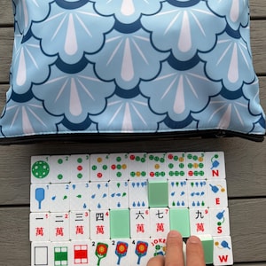 May include: A Mahjong set with white tiles featuring Chinese characters and colorful symbols. The set includes green tiles and is displayed on a wooden surface. A light blue zippered pouch with a scalloped pattern is in the background.